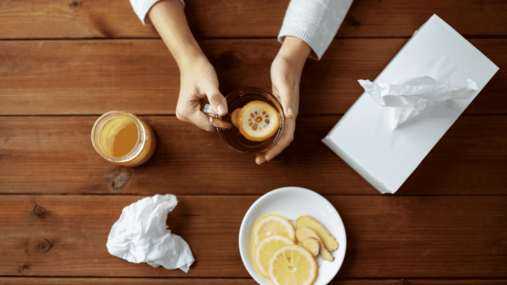 Ingredientes contra a gripe sobre uma mesa, além de lenços de papel e uma pessoa segurando uma xícara de chá