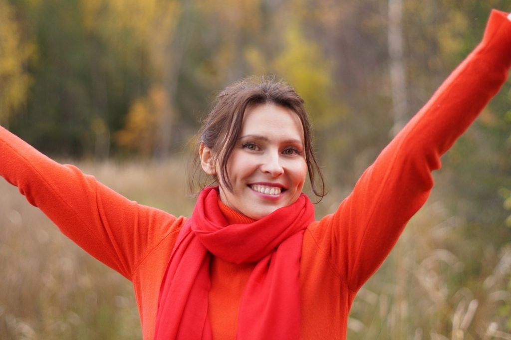 Imagem de uma mulher feliz. Ela está sorrindo e mostrando o quanto ela é resiliente, forte e corajosa mediante os seus problemas. Ela usa uma blusa de manga longa na cor laranja e um cachecol no pescoço na cor vermelha.