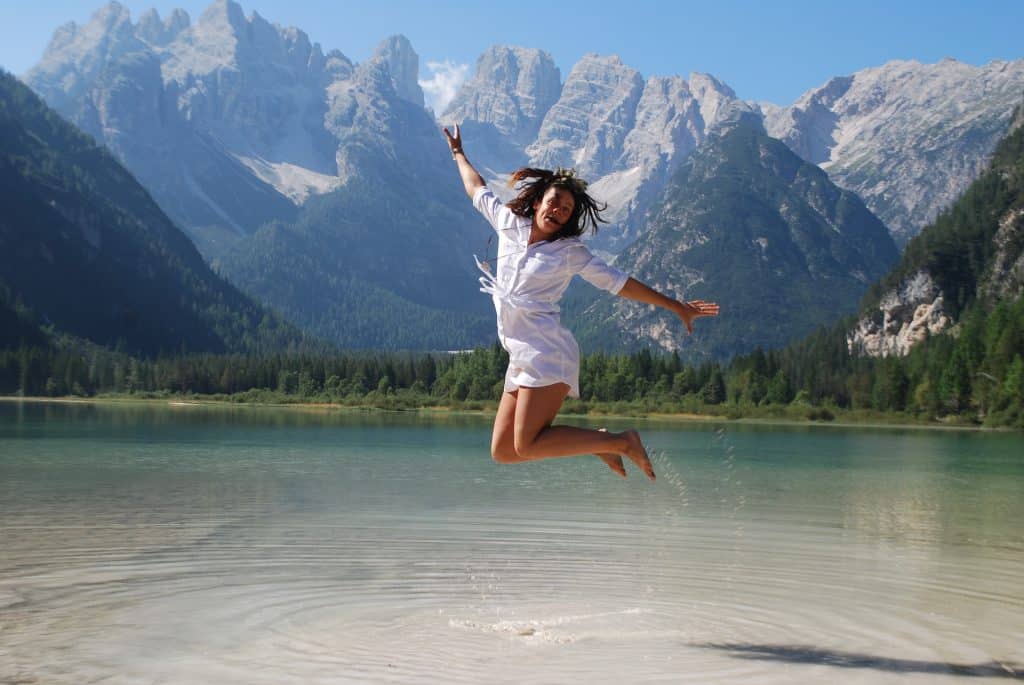 Imagem de uma jovem garota vestindo uma samisa e um shorts na cor branca. Ela está dando um salto em um lago.  Ao fundo algumas montanhas e uma floresta com vários pinheiros.