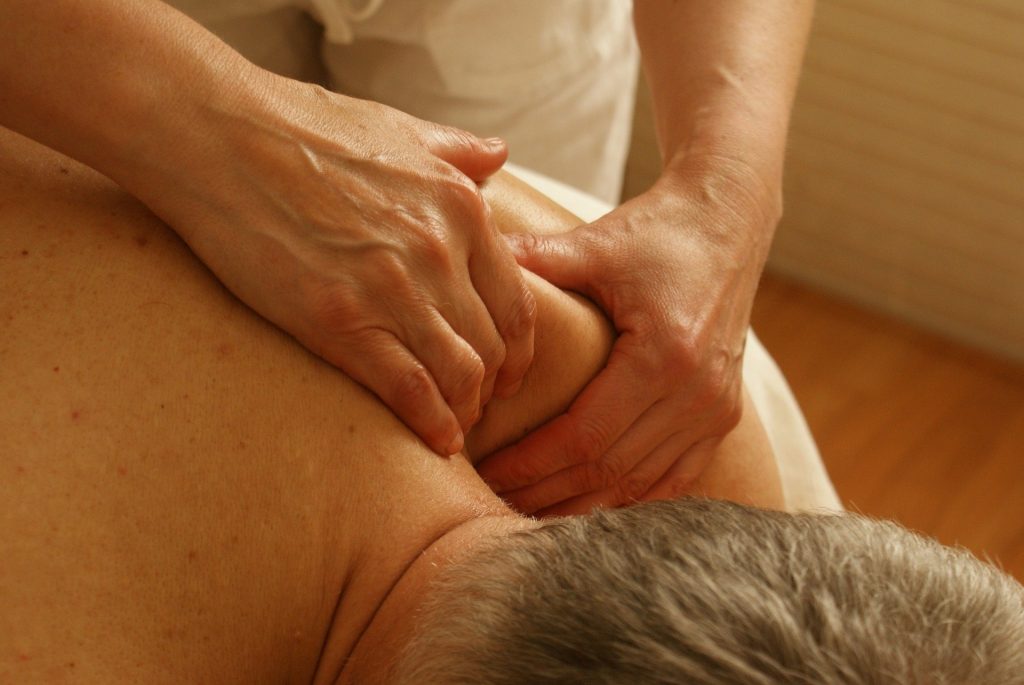 Imagem de um senhor de cabelos grisalhos deitado de costas sobre uma maca. Ele está sem camisa e recebe de  uma terapeuta uma sessão de quiropraxia em seu ombro.
