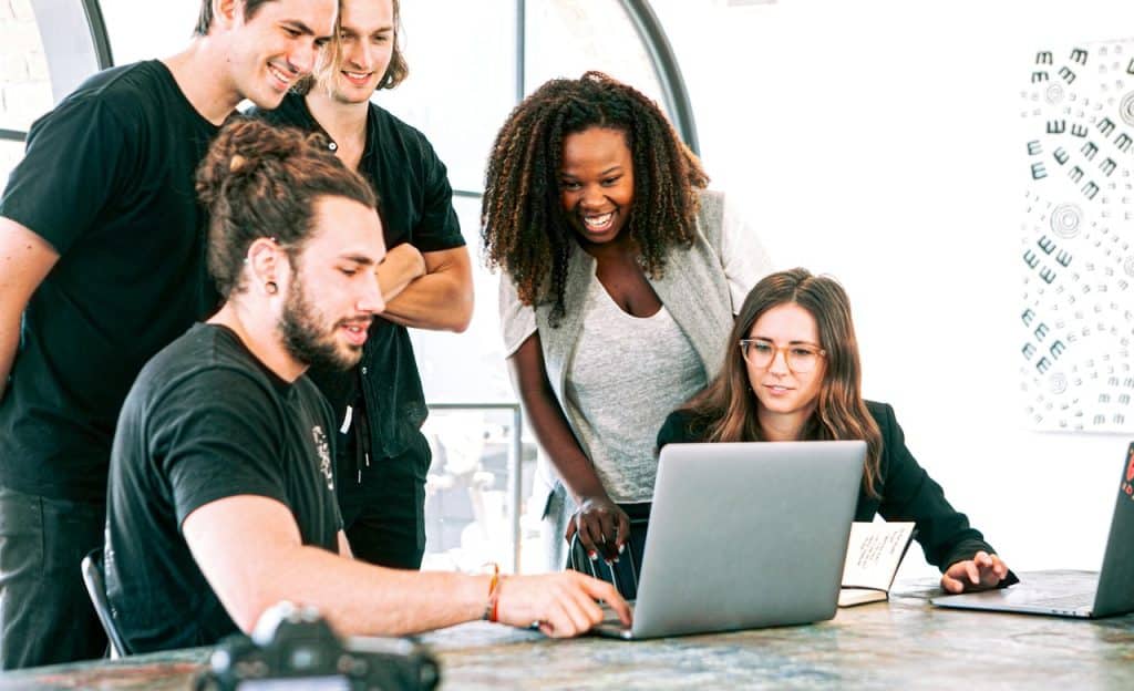 Grupo de cinco pessoas olham computador que está sobre uma mesa. Duas delas, um homem e uma mulher, estão sentados à mesa.