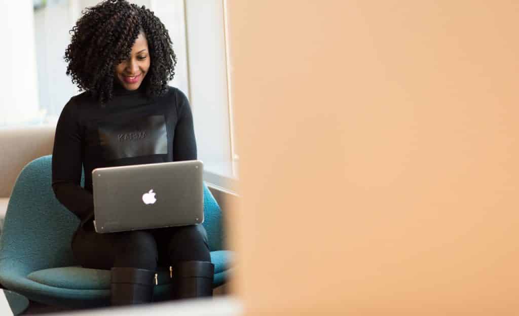 Mulher sentada com um computador sobre o colo. Ela olha para a tela do aparelho e sorri.