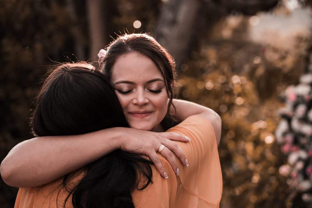 Duas mulheres se abraçando. Uma delas usa uma grande aliança de ouro em seu dedo.