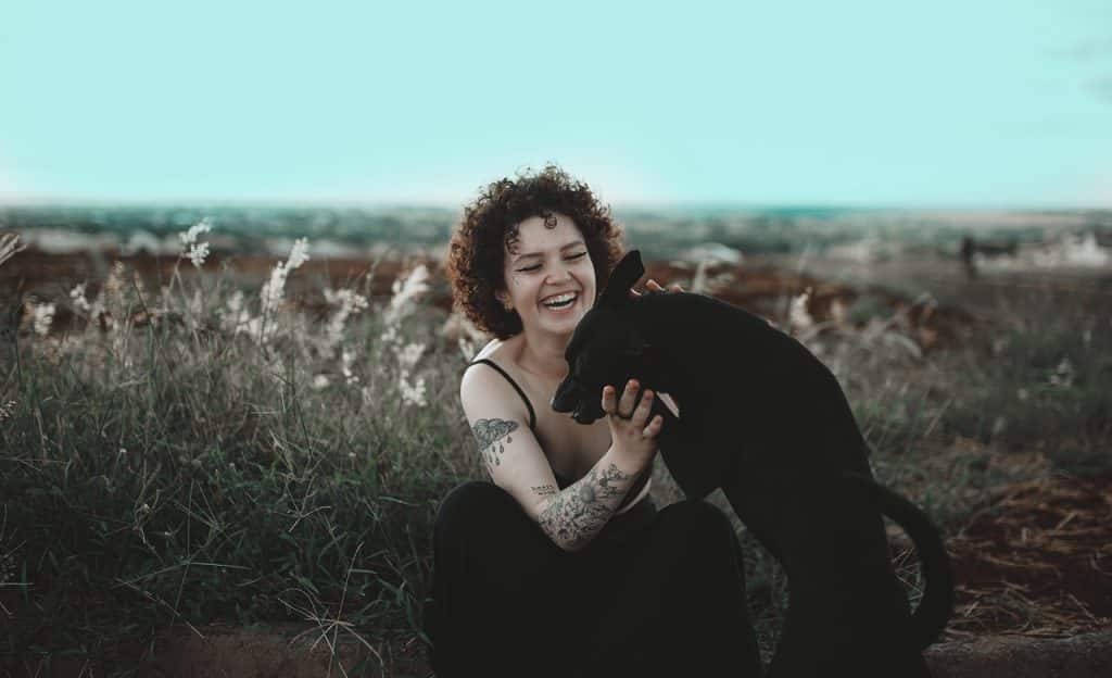 Em cenário externo, mulher sorri e brinca com cachorro preto. 