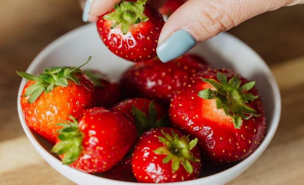 Mão feminina pega um morango do interior de uma tigela branca, onde há outros morangos.