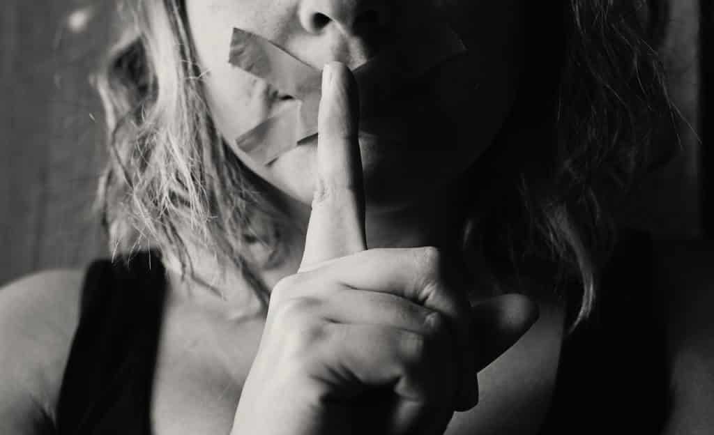 Esparadrapo sela boca de mulher. Seu dedo indicativo está sobre os lábios vedados, indicando silêncio.