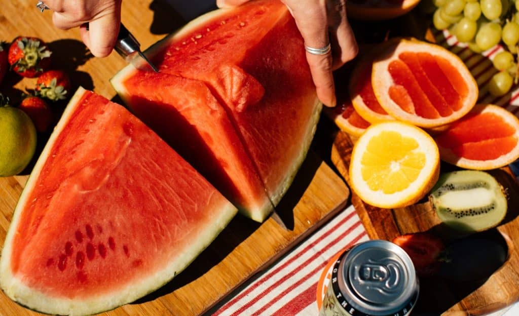 Mão corta melancia que está sobre mesa de madeira. Ao lado, também há outras frutas e uma lata fechada.