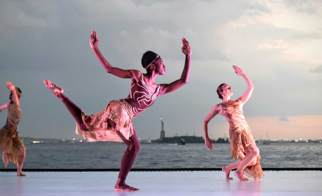 Três mulheres dançam em superfície próxima de um lago. 