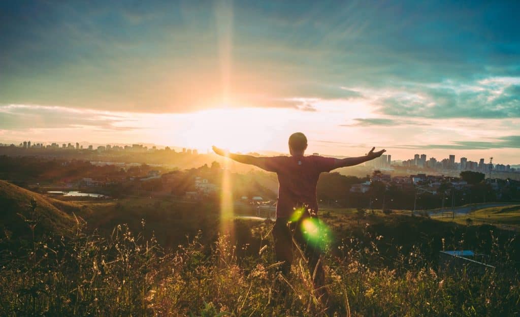 Homem com os braços abertos observa cidade de um morro. 