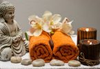 Imagem de um kit para uso na quiropraxia composto por duas toalhas na cor laranja, pedra, flores brancas, velas e a estátua do Buda.