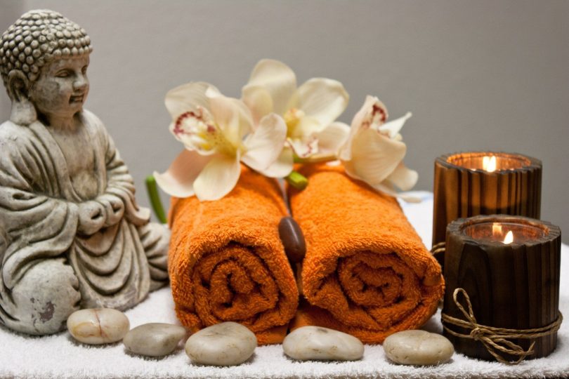 Imagem de um kit para uso na quiropraxia composto por duas toalhas na cor laranja, pedra, flores brancas, velas e a estátua do Buda.