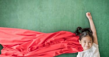 Garota deitada sobre cenário verde. Ela está com um dos braços erguidos e veste uma capa vermelha.