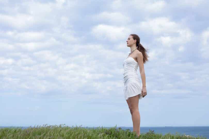 Mulher em ambiente externo. Ela está de pé sobre gramado e com os olhos fechados.