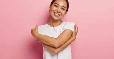 Mulher sorri e abraça a si mesma em fundo rosa.