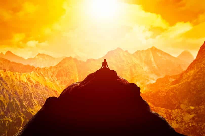 Mulher sentada no topo de uma montanha, ela está em prática meditativa. O ambiente é solar.