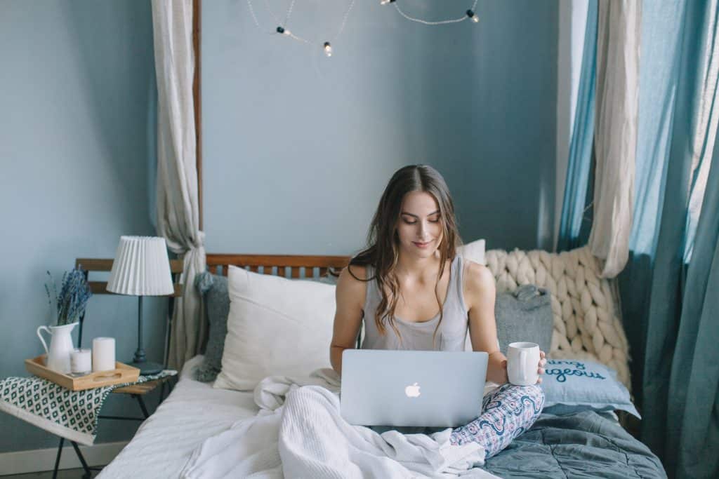 Mulher branca sentada numa cama com notebook no colo.