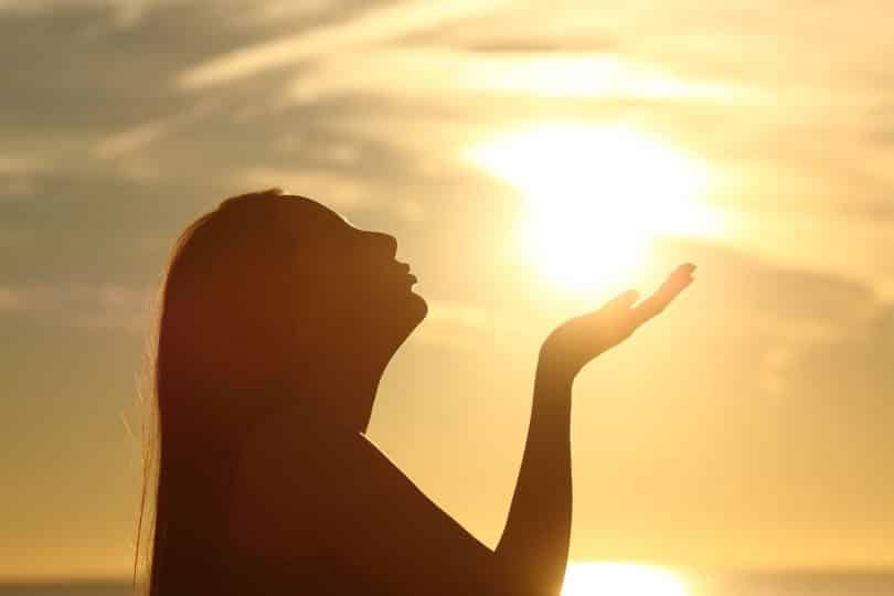 Mulher em ambiente externo ergue uma das mãos e inclina a cabeça para trás. Ao fundo, há o sol.