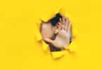 Face de uma mulher aparecendo parcialmente em um buraco feito em uma espécie de papel amarelo. Ela mostra sua orelha com a mão espalmada atrás dela, como se tentasse ouvir algo.