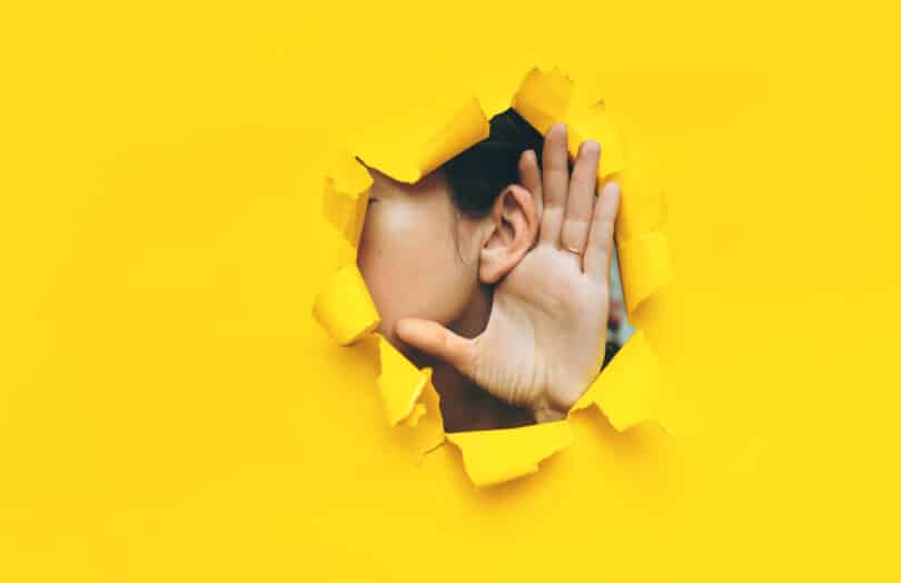 Face de uma mulher aparecendo parcialmente em um buraco feito em uma espécie de papel amarelo. Ela mostra sua orelha com a mão espalmada atrás dela, como se tentasse ouvir algo.