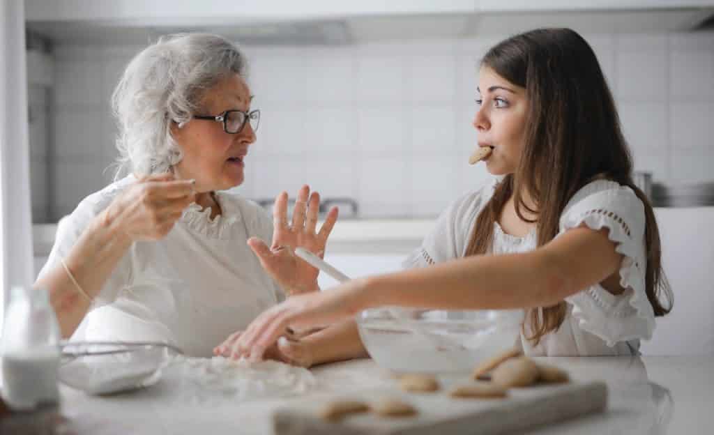 Uma menina e sua avó fazem biscoitos. As duas estão sentadas à bancada e olham uma para a outra. 