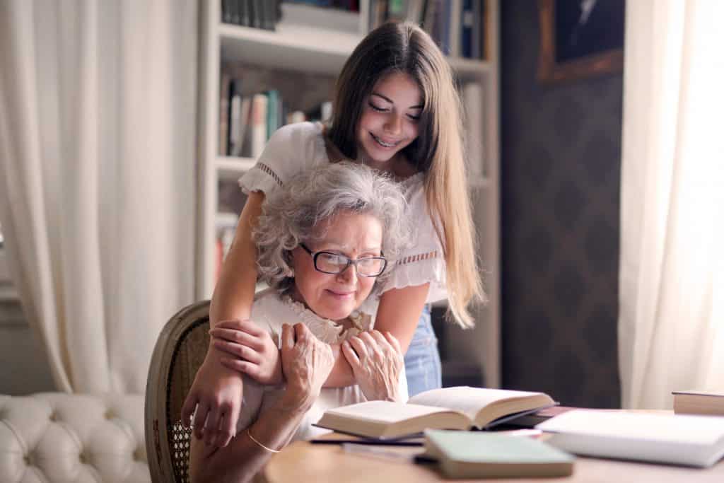 Menina abraça avó sentada à mesa; sobre esta, há um livro aberto para o qual ela olha.