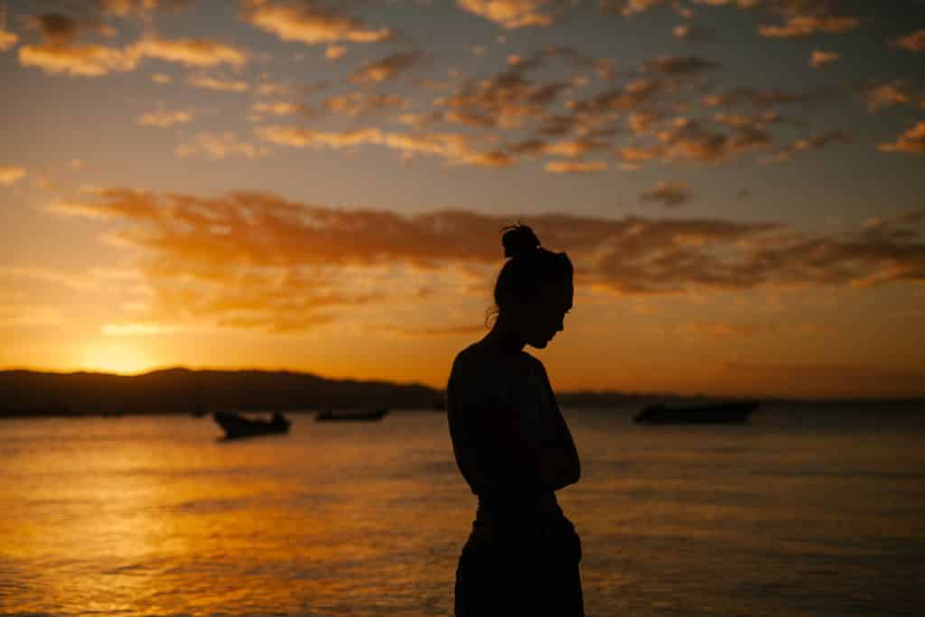 Mulher cabisbaixa em cenário externo. Há uma praia e o pôr do sol. 