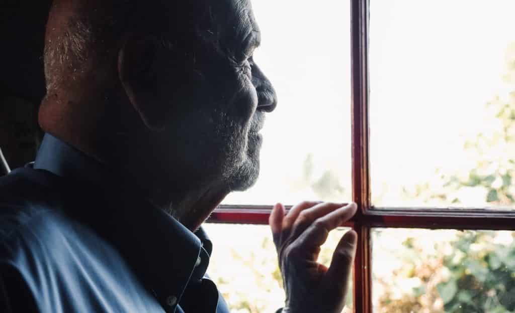 Homem idoso observa o ambiente externo através da janela. 