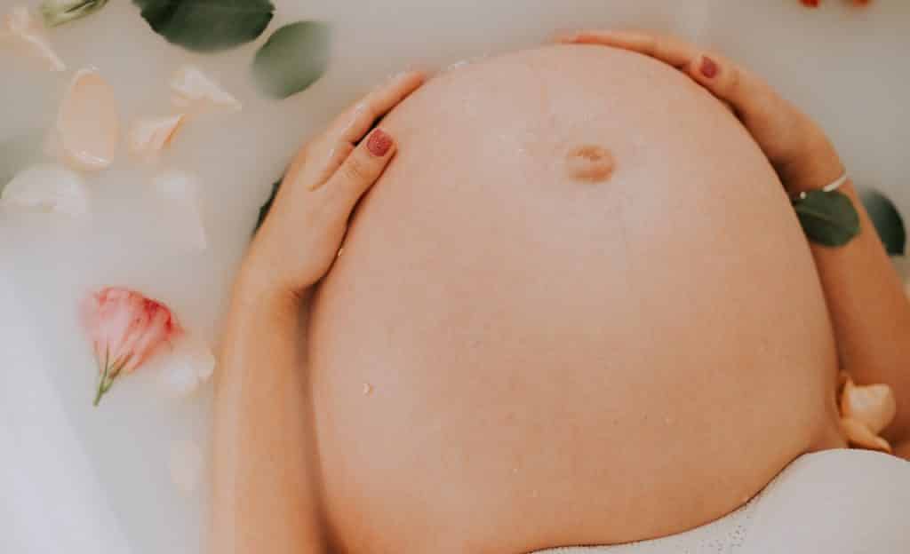 Mulher gestante toca o próprio abdômen. Ela está em uma banheira com espuma.