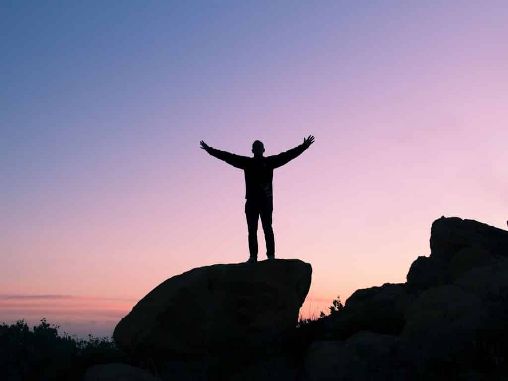 Homem em cima de uma pedra de abraços abertos durante o pôr do Sol