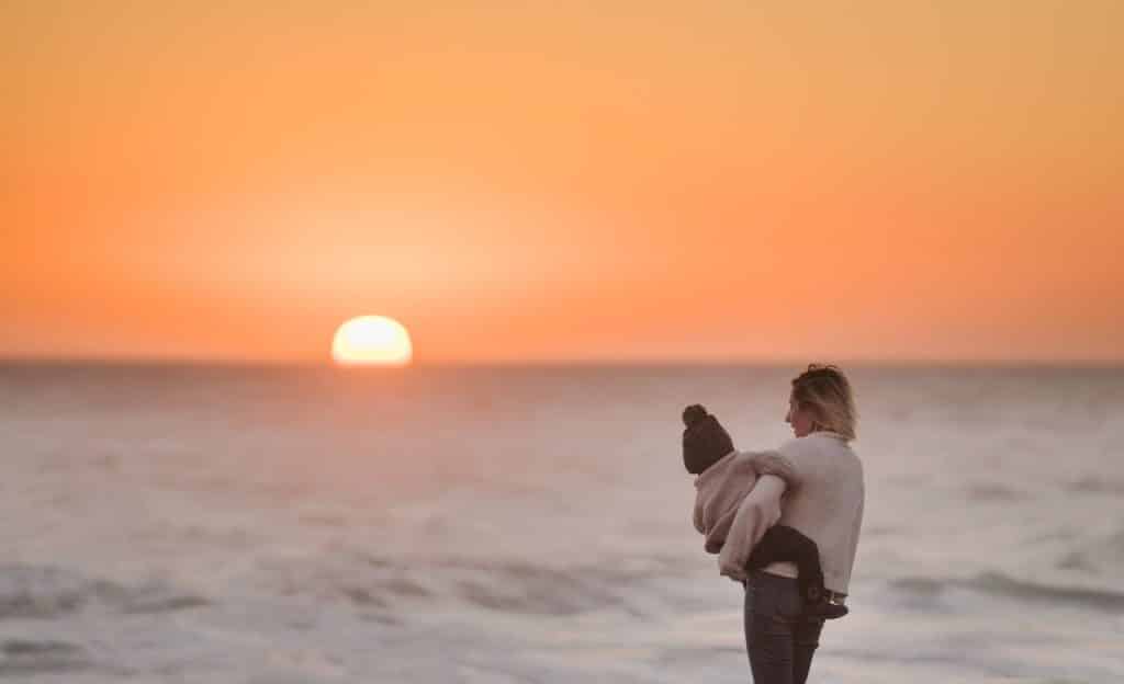 Mulher carrega criança. O cenário é de pôr do sol.