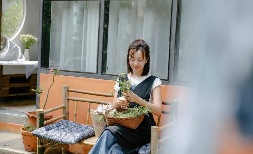 Mulher sentada em banco de madeira manipula plantas no interior de um cesto que está sobre o seu colo.