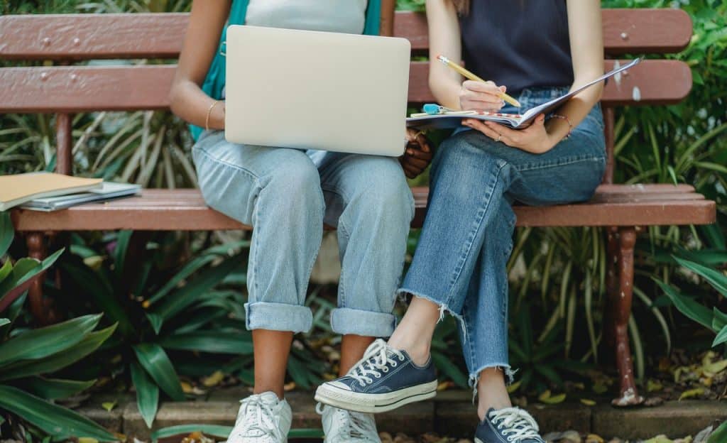 Duas mulheres estudam juntas em um banco de praça. Uma, à direita, está com um notebook sobre as pernas, enquanto a outra segura um caderno e escreve nele.