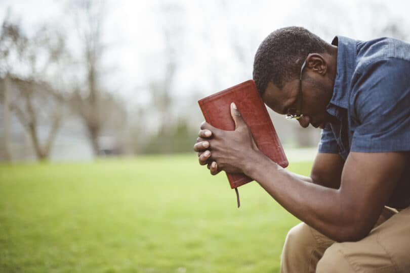 Homem negro sentado e segurando a Bíblia, de cabeça abaixada, como se estivesse refletindo ou rezando