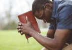 Homem negro sentado e segurando a Bíblia, de cabeça abaixada, como se estivesse refletindo ou rezando