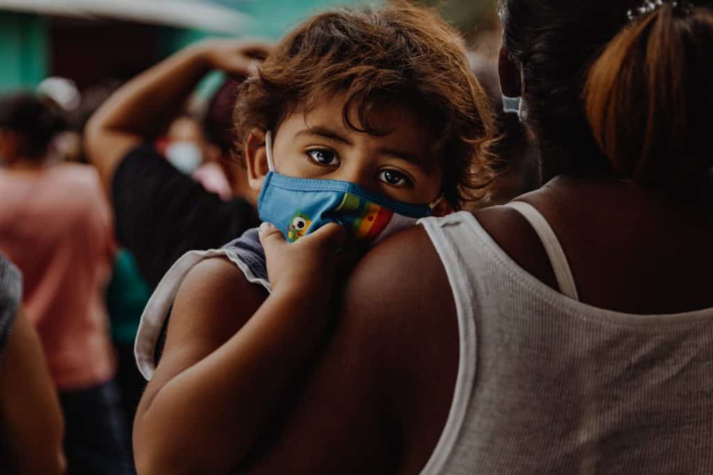 Menino negro de máscara no colo da mãe.