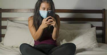 Mulher sentada em cama mexe em celular. Ela veste uma máscara azul no rosto.