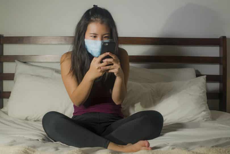 Mulher sentada em cama mexe em celular. Ela veste uma máscara azul no rosto.