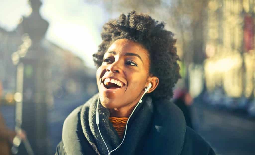 Mulher com fones de ouvido brancos sorri. Ela está em ambiente externo.