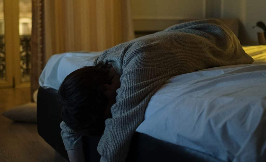Homem deitado de bruços em cama; ele está cabisbaixo.