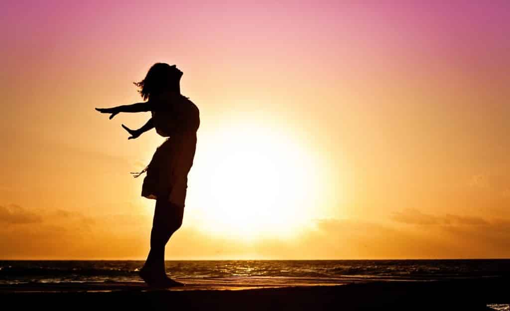 Mulher inclinada para trás com os braços abertos. Ela está em praia. Ao fundo, há o sol.