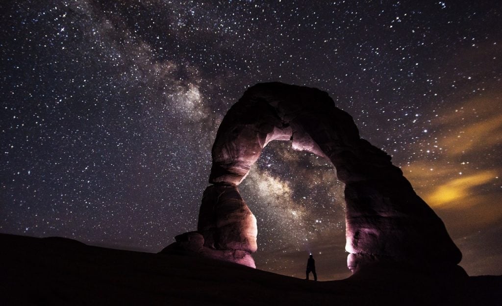 Pessoa sob arco de pedra à noite. O céu é estrelado.