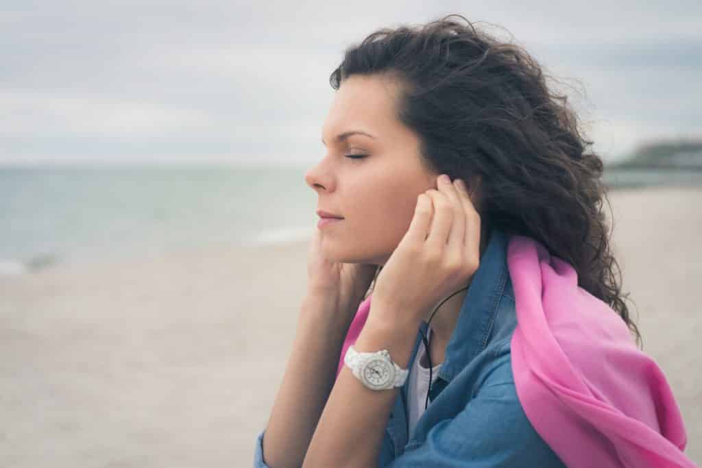 Mulher branca na praia com os olhos fechados e fones de ouvido.