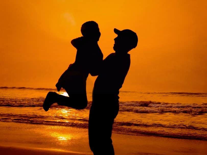 Silhueta de homem segurando criança numa praia.