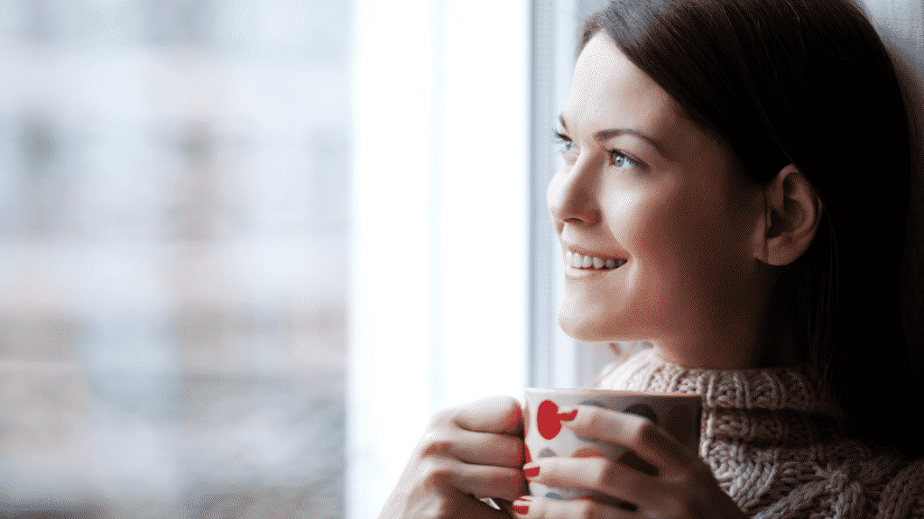 Mulher sorridente olhando pela janela segurando xícara de café