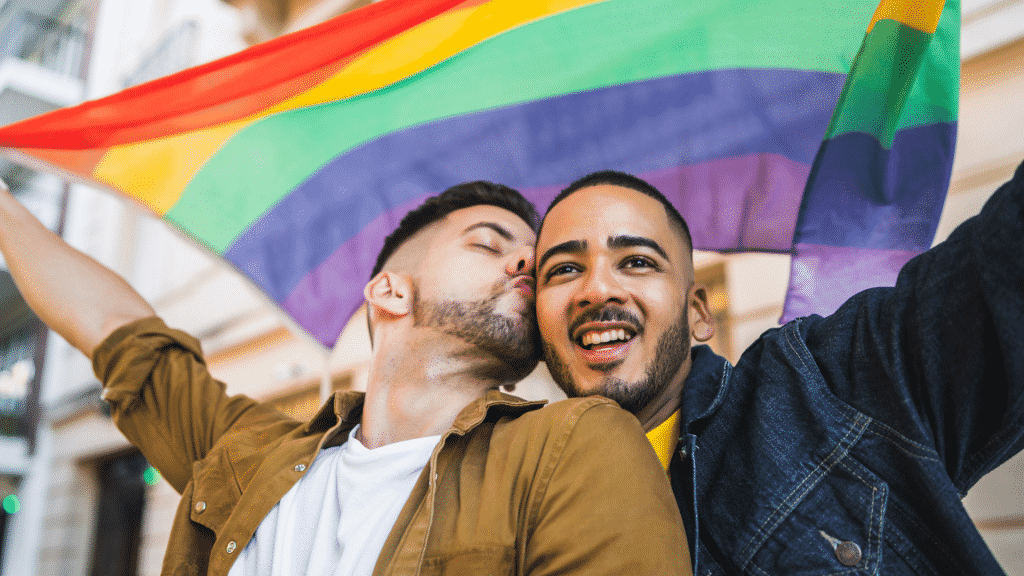 Casal gay segurando a bandeira LGBTQIA+. Um deles está beijando o parceiro no rosto