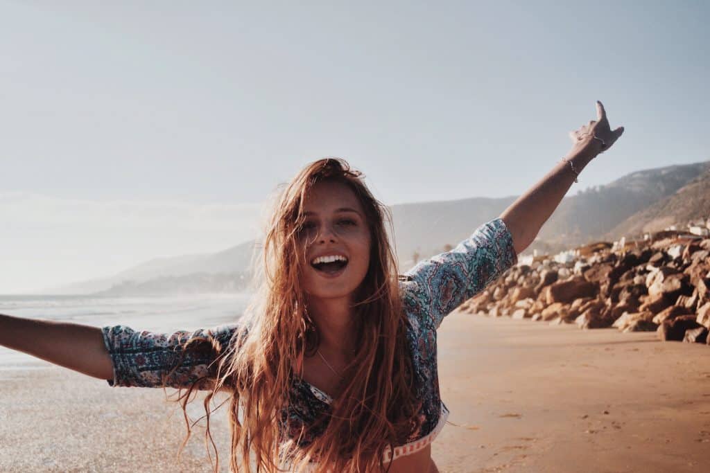 Mulher branca na praia com os braços abertos sorrindo.