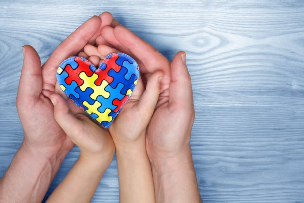 Mãos de um adulto e de uma criança segurando o símbolo do autismo - um coração com um quebra-cabeça dentro