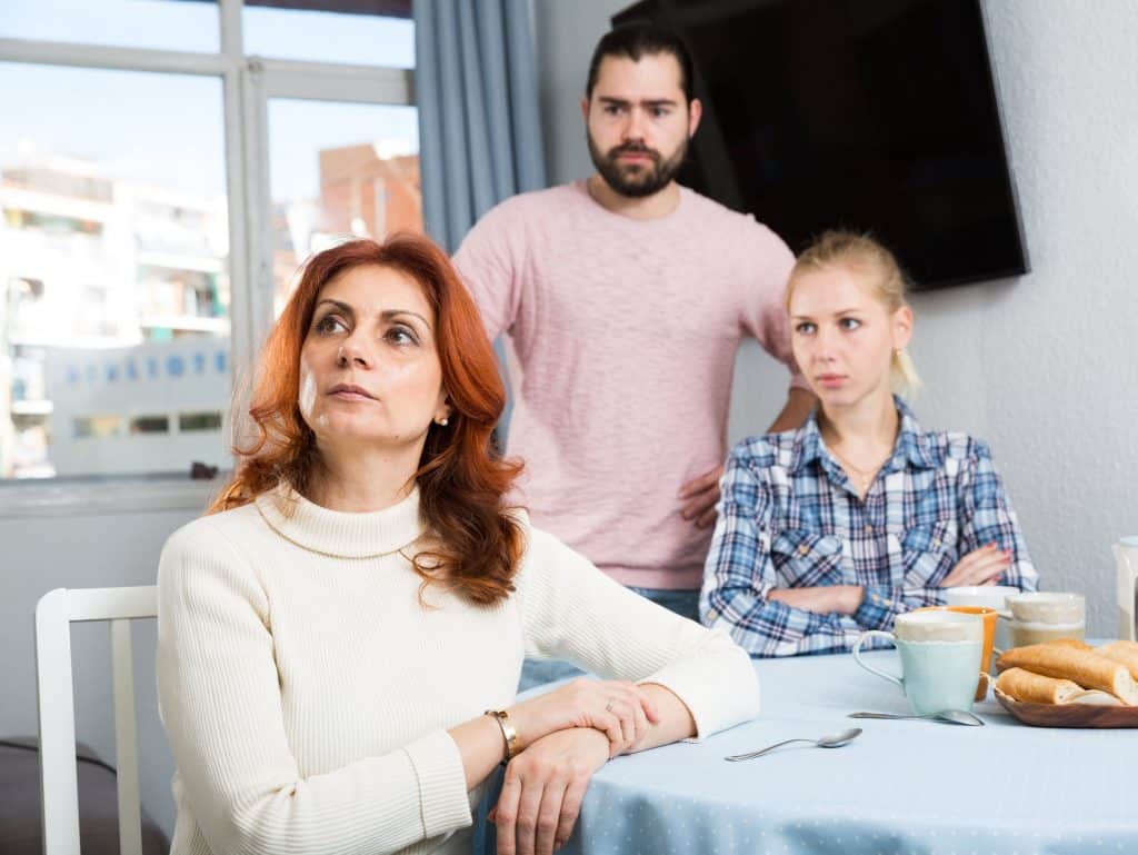 Uma mulher sentada à mesa olhando pra cima. Ao fundo, uma mulher e um homem. Todos eles com semblante de evidente desconforto.