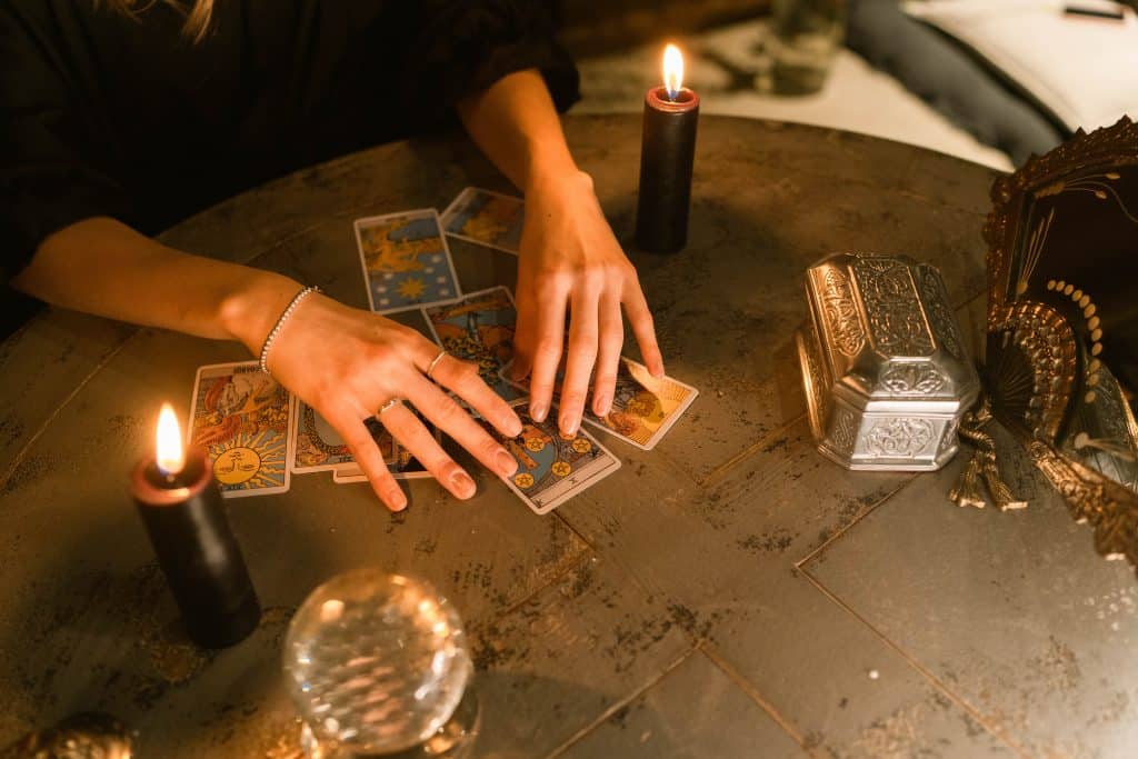 Mãos de uma mulher manipulando um baralho de cartas. Sobre uma mesa, uma vela preta acesa e um leque de ferro, além de um globo cinza e também uma caixa acinzentada. 