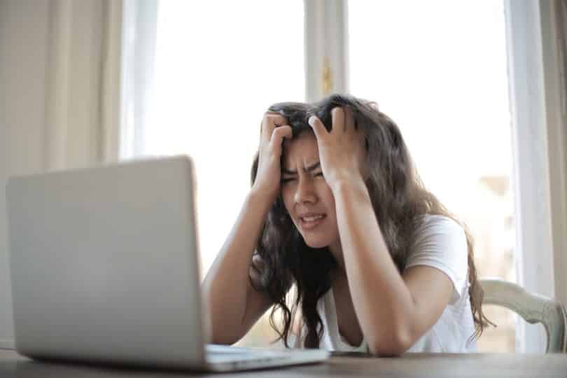 Uma mulher com as mãos sobre a cabeça, exibindo um semblante de estresse. Na sua frente, um notebook.